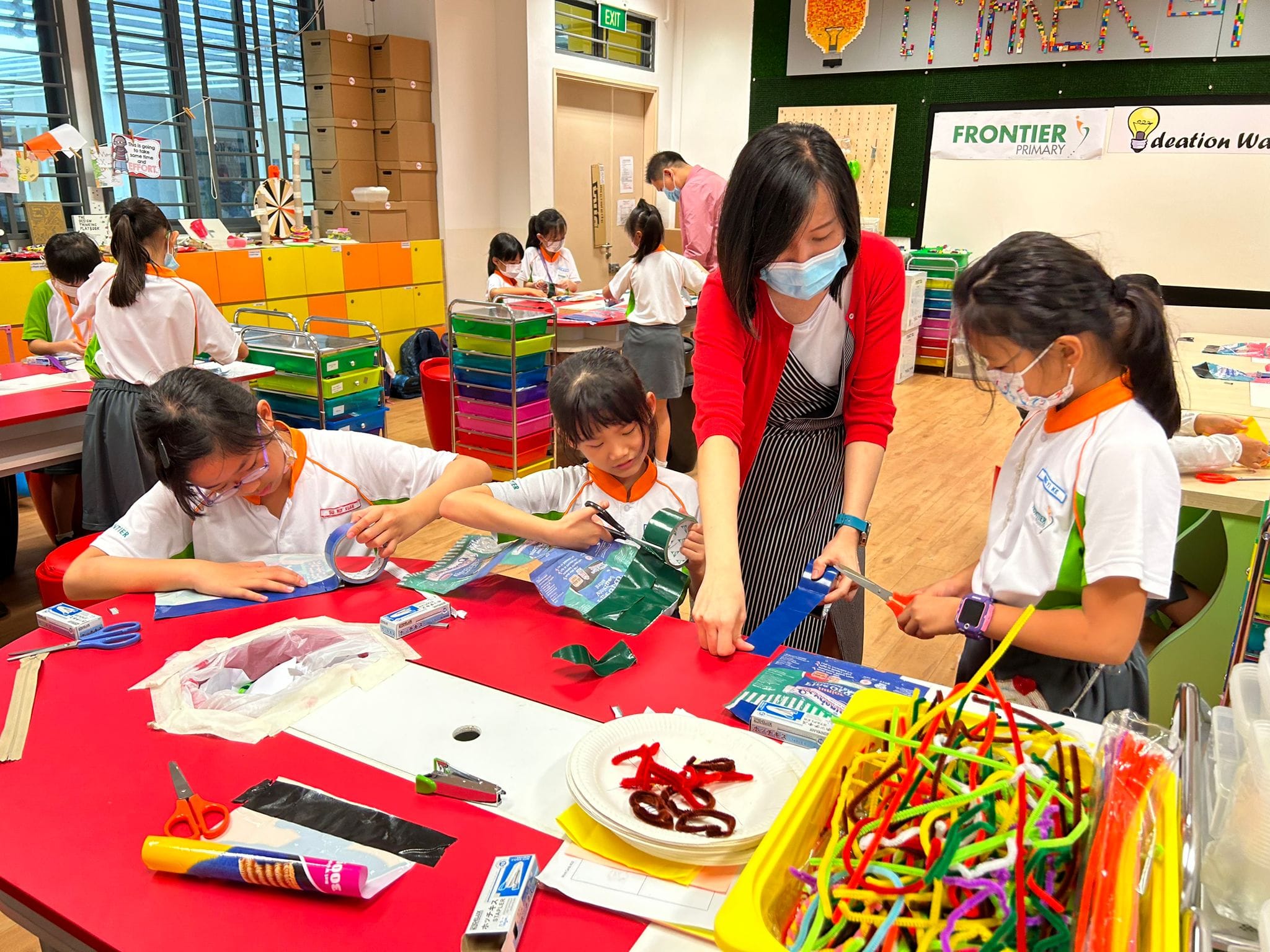 A masked teacher helps young students with a craft project at a colorful table in Frontier Primary school's ideation room.